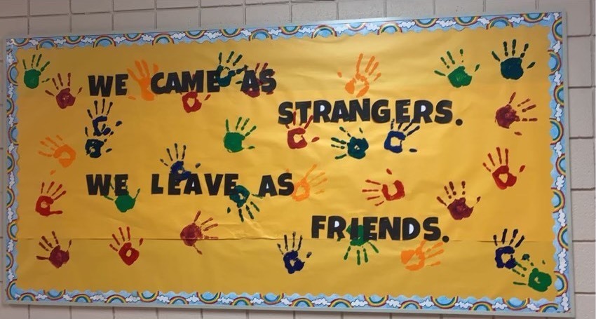 Yellow bulletin board with rainbow borders and multi-colored handprints with the text "We came as strangers.  We leave as friends."