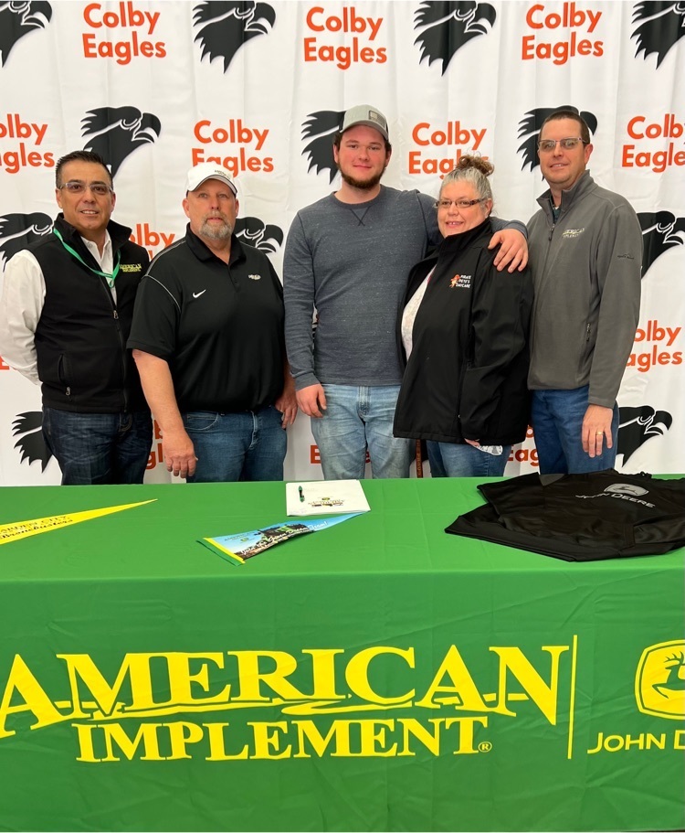 A high schooler, mom, and American Implement representatives behind an American Implement tablecloth and in front of Colby Eagles background.